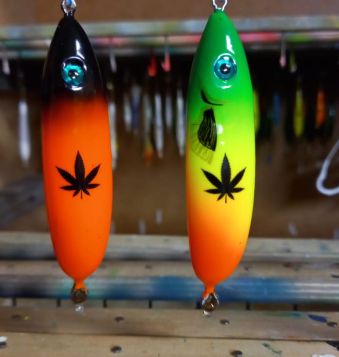 Two handmade lures in the workshop: a black one with an orange gradient and a green one with yellow, each with a black leaf pattern printed on it
