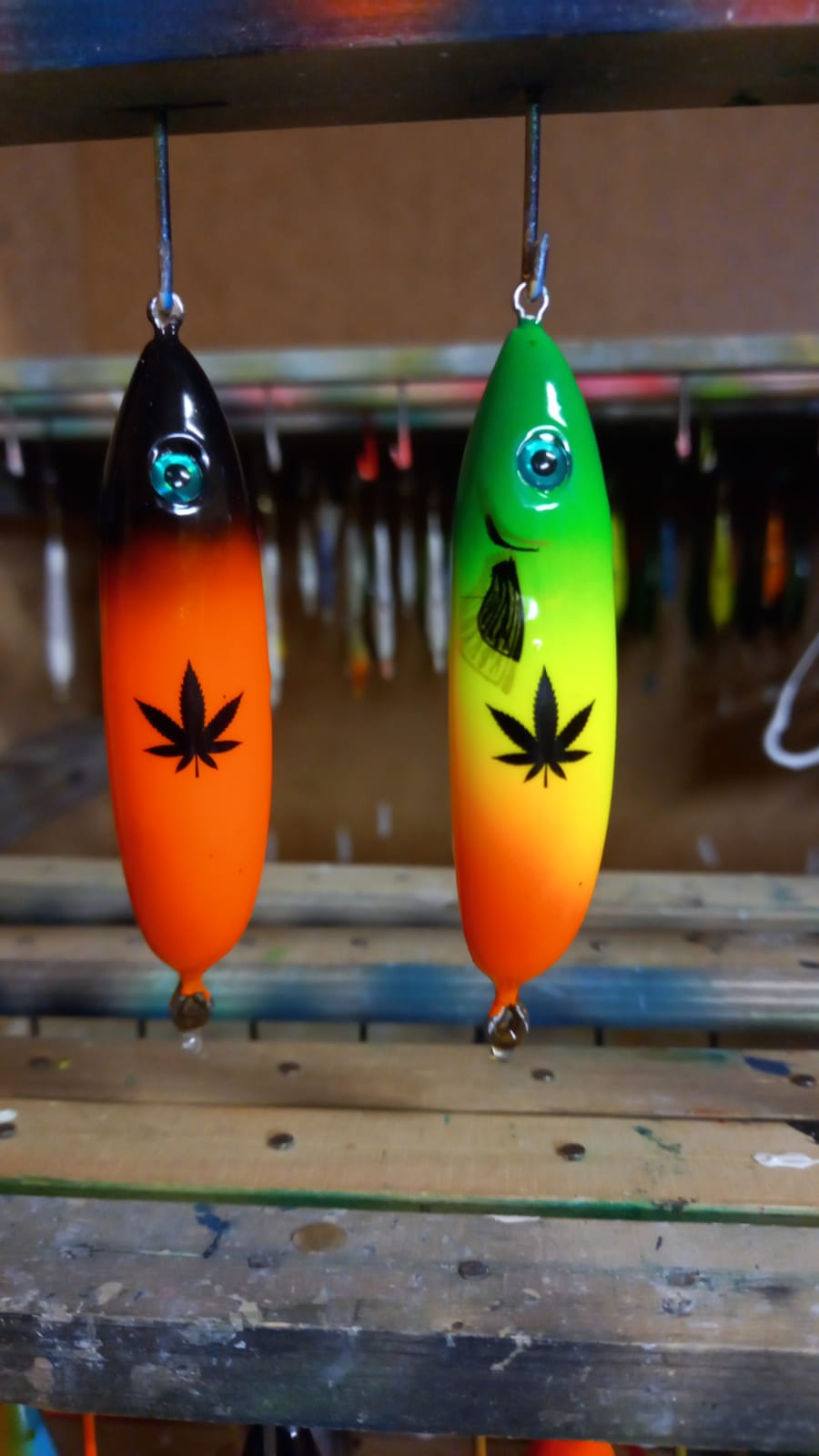 Two handmade lures in the workshop: a black one with an orange gradient and a green one with yellow, each with a black leaf pattern printed on it
