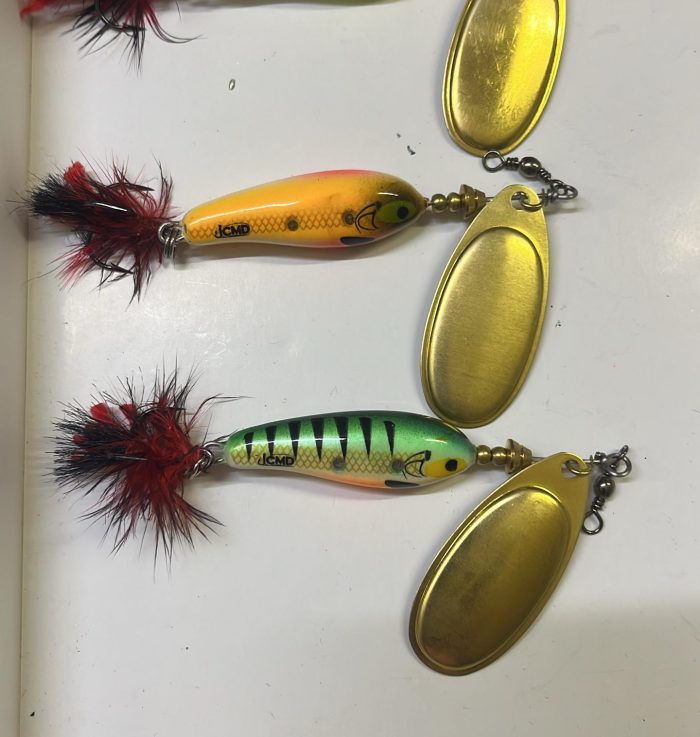 Three handmade spinner baits with golden blades and red-and-black tail feathers on a white surface: bluish-yellow, orange and green with black stripes