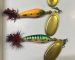 Three handmade spinner baits with golden blades and red-and-black tail feathers on a white surface: bluish-yellow, orange and green with black stripes