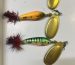 Three handmade spinner baits with golden blades and red-and-black tail feathers on a white surface: bluish-yellow, orange and green with black stripes