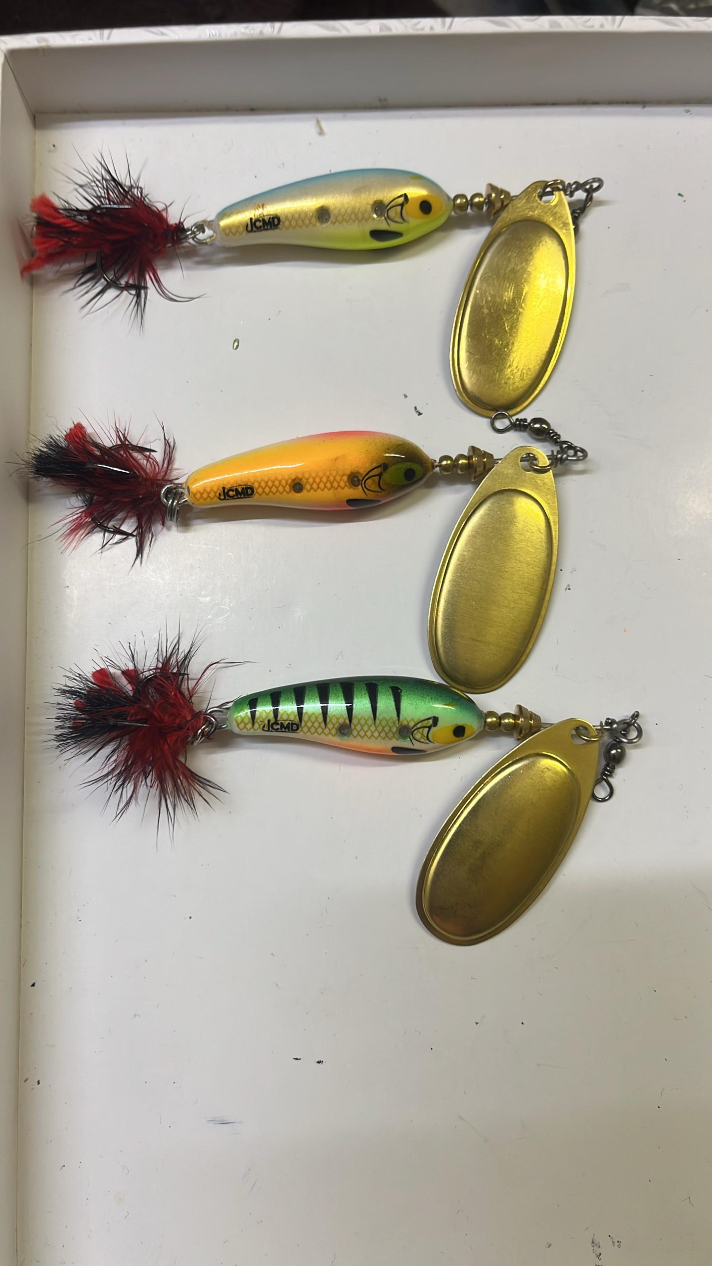 Three handmade spinner baits with golden blades and red-and-black tail feathers on a white surface: bluish-yellow, orange and green with black stripes